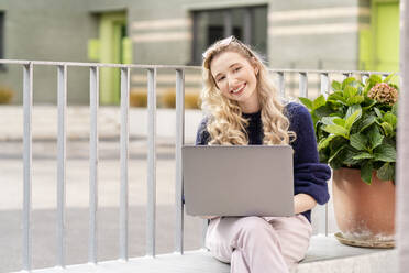 Happy young blond woman with laptop sitting on wall ledger - PESF03514