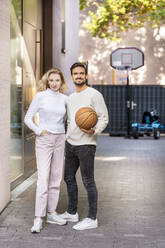 Young man holding basketball standing with girlfriend on footpath - PESF03519