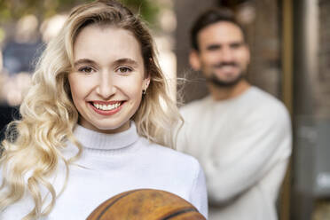 Happy blond woman with basketball standing in front of boyfriend - PESF03521