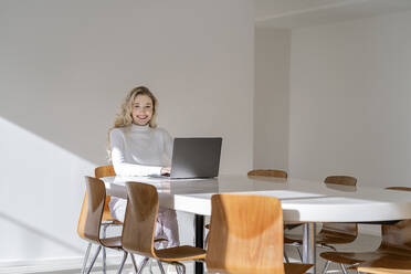 Glückliche blonde Frau mit Laptop sitzt am Tisch - PESF03525