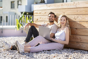 Glückliches junges Paar sitzt mit Laptop an einem sonnigen Tag - PESF03534