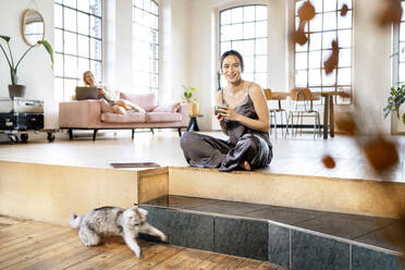 Smiling businesswoman with coffee cup sitting cross-legged at home - DMOF00192