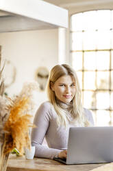 Smiling blond businesswoman with laptop sitting at home - DMOF00197