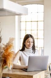 Smiling young woman working on laptop at home - DMOF00198