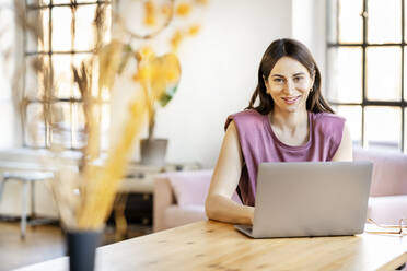Lächelnde schöne Geschäftsfrau mit Laptop sitzt im Wohnzimmer zu Hause - DMOF00215