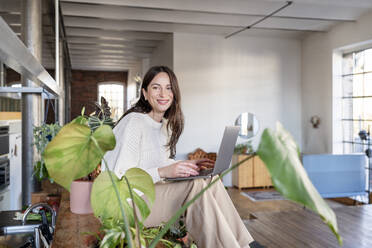 Smiling businesswoman with laptop sitting on table at home - DMOF00225