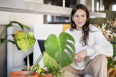 Glückliche Frau sitzt bei Topfpflanzen zu Hause - DMOF00229