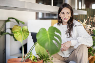 Happy woman sitting by potted plants at home - DMOF00229