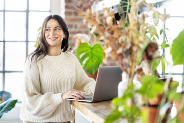Lächelnde Geschäftsfrau, die mit ihrem Laptop zu Hause neben Topfpflanzen steht - DMOF00231
