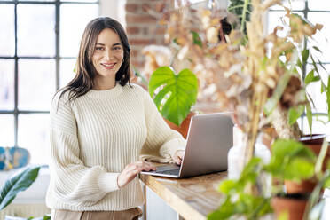 Happy businesswoman with laptop standing by potted plants in living room at home - DMOF00232