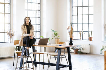 Businesswoman wearing headset sitting by laptop on table at home office - DMOF00274