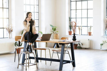 Businesswoman wearing headset sitting on table at home office - DMOF00275