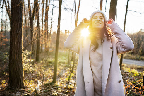 Glückliche Frau mit Strickmütze, die an einem sonnigen Tag bei Bäumen im Wald steht - DMOF00298