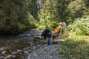 USA, Idaho, Sun Valley, Frau sitzt auf Stuhl am Flussufer im Wald - TETF00621