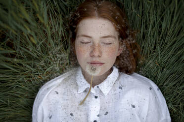 Teenager-Mädchen mit roten Haaren und Sommersprossen liegt im Gras und hat eine Pusteblume an den Lippen. Ihre Augen sind geschlossen. - TETF00659