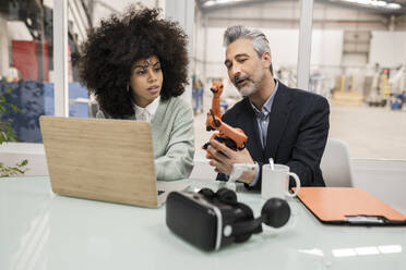 Geschäftsmann erklärt einem Kollegen, der in einer Fabrik am Schreibtisch sitzt, einen Roboterarm - JCCMF05524