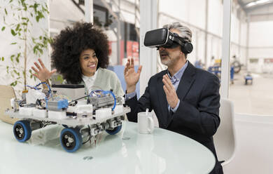 Ein lächelnder Entwickler betrachtet einen Geschäftsmann, der einen Virtual-Reality-Simulator in einer Fabrik trägt - JCCMF05528