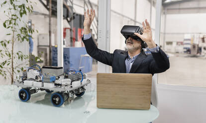 Businessman wearing virtual reality sitting with hands raised in factory - JCCMF05532