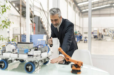 Businessman holding coffee cup examining robotic vehicle model standing at desk - JCCMF05576