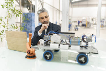 Businessman sitting with hand on chin looking at model of robotic vehicle in factory - JCCMF05579