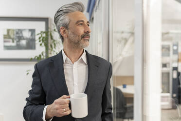 Businessman holding coffee cup standing glass wall in factory - JCCMF05581