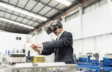 Geschäftsmann mit grauem Haar, der virtuelle Realität trägt und in einer Fabrik gestikuliert - JCCMF05602