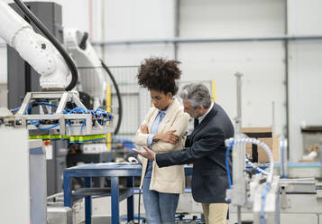 Engineer explaining robotic arm to developer standing with arms crossed in factory - JCCMF05630