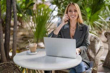 Smiling blond businesswoman with laptop talking on smart phone sitting at sidewalk cafe - DLTSF02774