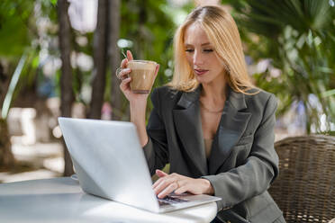 Blonde Freiberuflerin mit Kaffeetasse und Laptop in einem Straßencafé - DLTSF02775