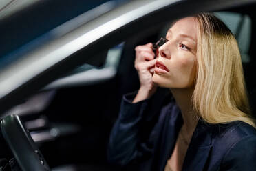 Blond woman applying mascara sitting in car - DLTSF02777