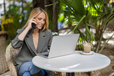 Smiling blond businesswoman talking on mobile phone using laptop sitting at sidewalk cafe - DLTSF02779