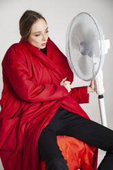 Junge Frau in rotem Mantel mit Blick auf einen elektrischen Ventilator vor weißem Hintergrund - IYNF00077