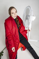 Schöne Frau in rotem Mantel sitzend mit elektrischem Ventilator vor weißem Hintergrund - IYNF00079