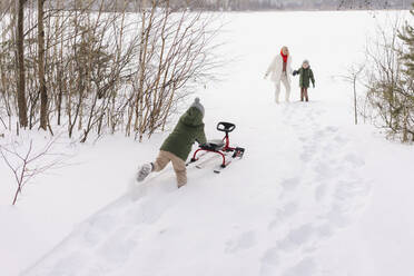 Frau mit Sohn betrachtet Junge, der im Winter Schlitten auf Schnee schiebt - SEAF00660