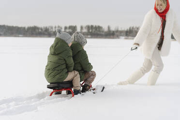 Boys sitting on toboggan pulled by mother walking on snow in winter - SEAF00671