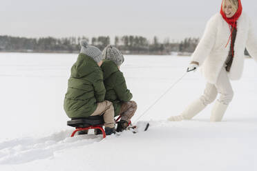 Jungen sitzen auf einem Schlitten, der von der Mutter gezogen wird, und gehen im Winter durch den Schnee - SEAF00671
