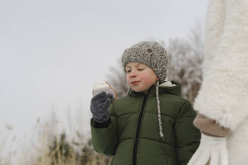 Boy wearing knit hat eating cookie standing by mother in winter - SEAF00678
