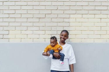 Happy young woman carrying daughter standing in front of wall - PGF01024