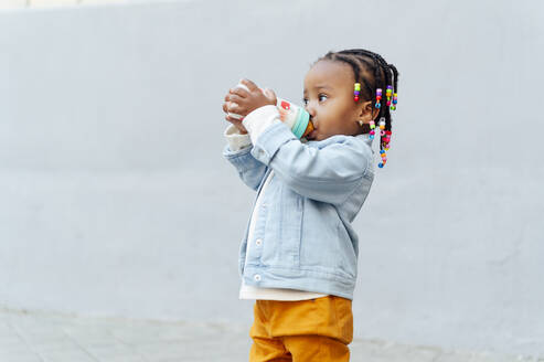 Cute little girl drinking milk from baby bottle in front of wall - PGF01025