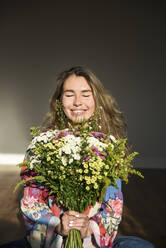 Lächelnde Frau mit geschlossenen Augen, die einen Blumenstrauß hält und zu Hause sitzt - VBUF00061