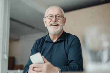 Glücklich arbeitender Mann mit Brille, der ein Mobiltelefon im Büro hält - JOSEF07339