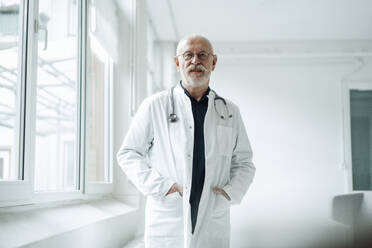 Doctor standing with hands in pockets by window at hospital - JOSEF07344