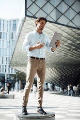 Smiling businessman using tablet PC standing on seat outside modern building - GUSF07137