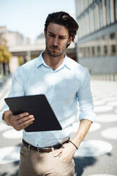 Geschäftsmann mit Hand in der Tasche, der an einem sonnigen Tag einen Tablet-PC benutzt - GUSF07138