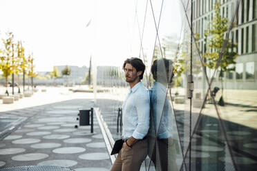 Businessman holding tablet PC standing with hand in pocket leaning on glass wall - GUSF07152