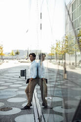 Businessman holding tablet PC standing with hand in pocket leaning on glass wall at modern building - GUSF07153