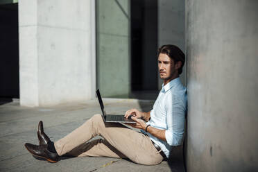 Businessman with laptop sitting by wall on sunny day - GUSF07162