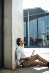 Businessman with laptop looking up sitting by by wall outside building - GUSF07164