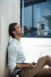 Handsome businessman with laptop looking up sitting by wall - GUSF07166