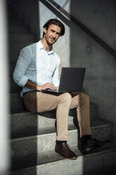 Smiling handsome businessman sitting with laptop on steps - GUSF07179